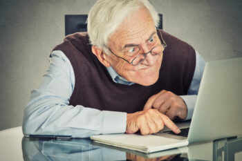 Elderly old man using computer sitting at table isolated on grey wall background. Senior people and technology concept